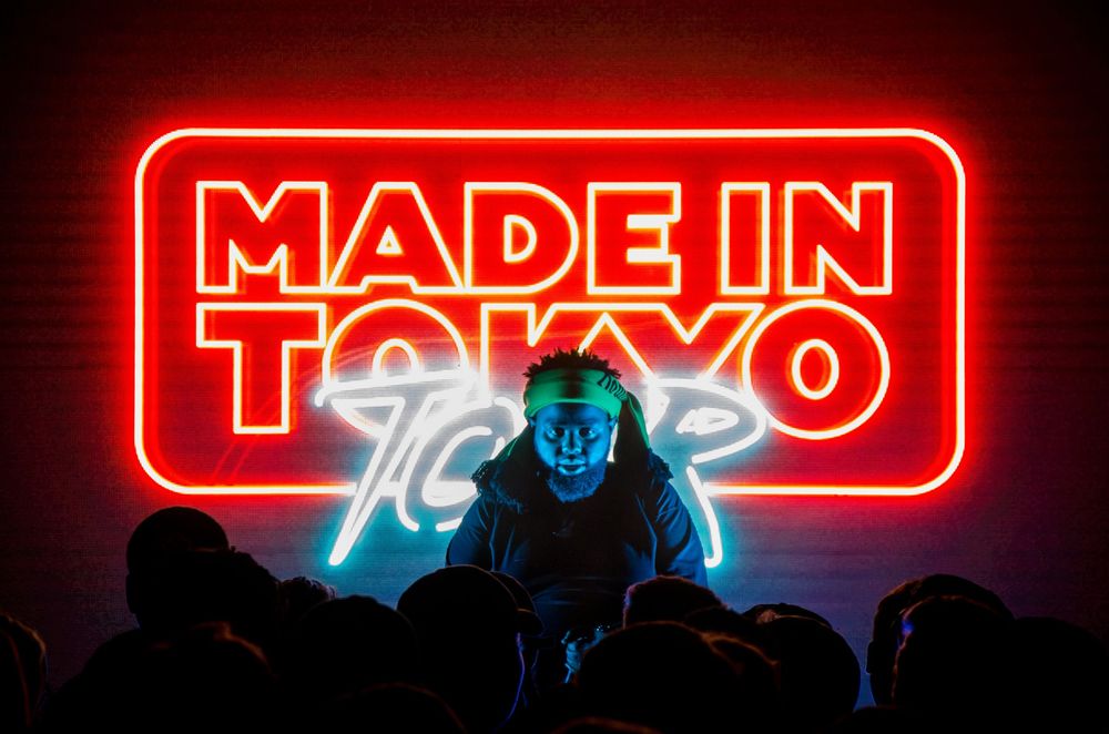 Performer on stage beneath a vibrant red neon sign reading Made In Tokyo Tour in a dark concert venue.