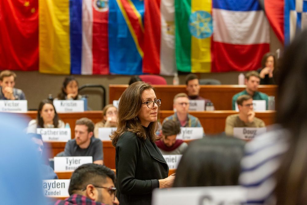 Woman in black outfit speaks at international conference with colorful country flags displayed in background.