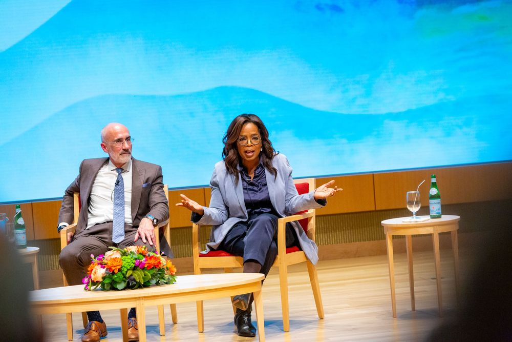 Two professionals engage in a panel discussion on stage, with a woman gesturing expressively beside a seated man.