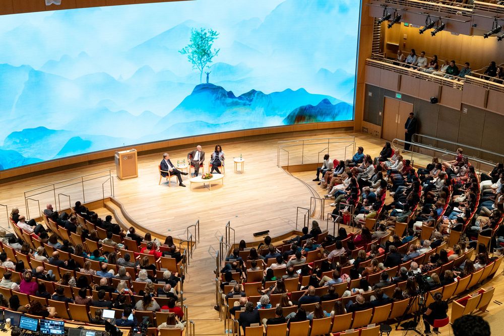 Panel discussion on stage in modern auditorium with blue mountain backdrop and tree centerpiece before large audience.
