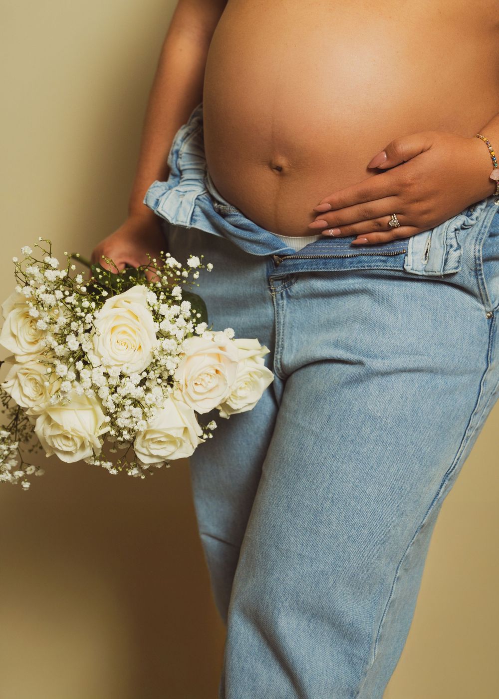 Pregnant woman in open denim jeans holding white roses bouquet, showcasing baby bump, maternity photography.