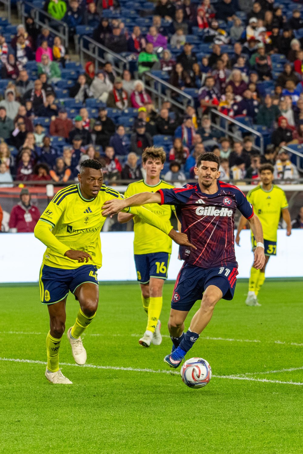 MLS soccer players compete for the ball during a night match, with one player in a red New England Revolution jersey.