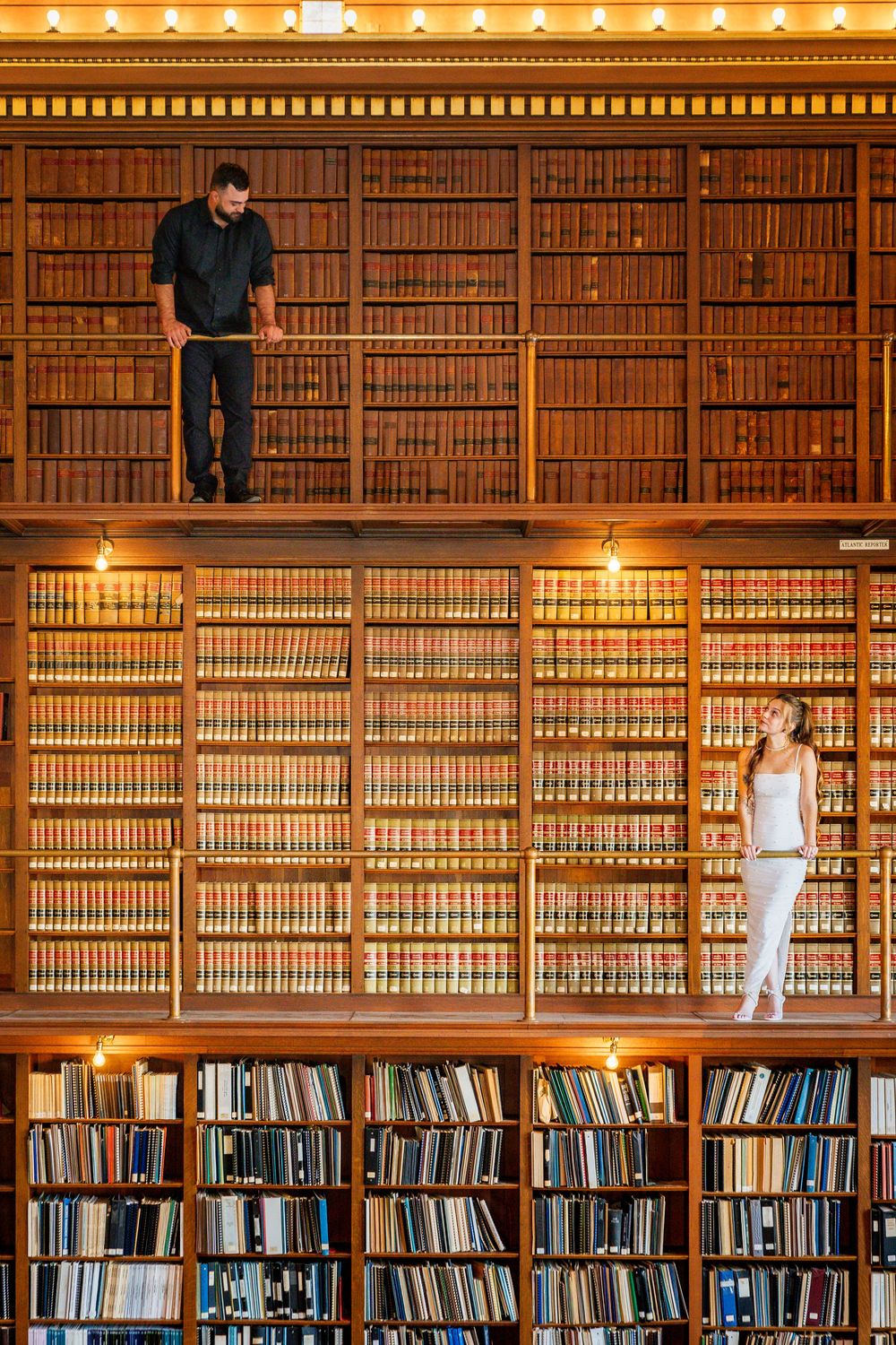 Man and woman standing on different levels of a grand wooden library filled with rows of books.
