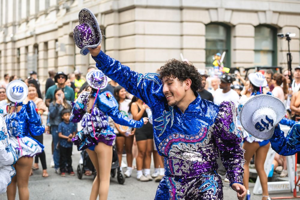 Performer in blue sequin costume waves at a vibrant street parade with dancers in matching outfits.