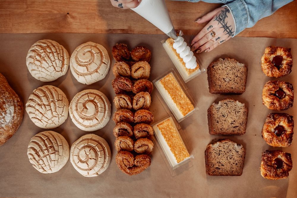 Assorted artisan baked goods displayed on a wooden table, including bread rolls, pastries, loaf slices, and glazed donuts.