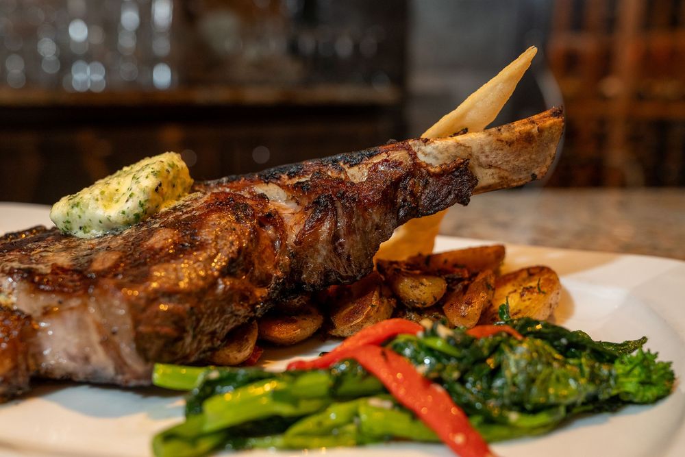 Grilled bone-in steak with herb butter, roasted potatoes, sautéed greens, and red chili on a white plate.