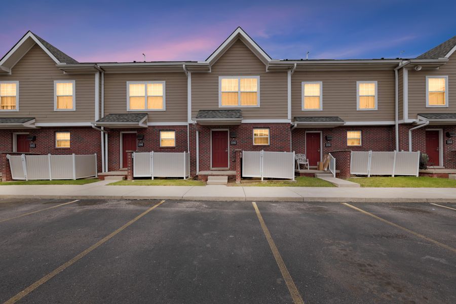 Twilight view of brick and siding townhomes with white fences, lit windows, and a parking lot in a residential complex.