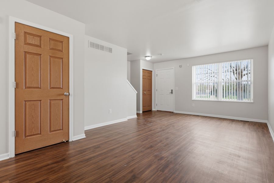 Bright empty living room with dark hardwood floors, white walls, a wooden door, and a window letting in natural light.
