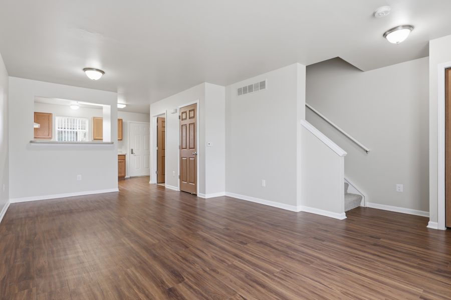 Spacious open living room with hardwood floors, white walls, staircase, and view into kitchen area.