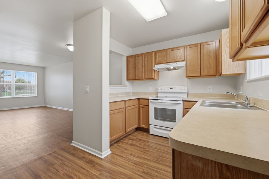 Modern apartment kitchen with oak cabinets, white appliances, and hardwood floors opening to a bright living area.