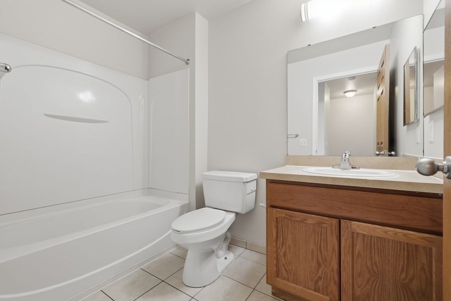 Clean full bathroom featuring a white soaking tub, toilet, and oak vanity with mirror on white tile floors.