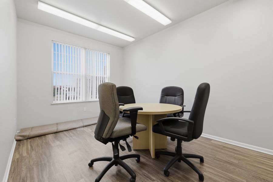 Small office meeting room with round wooden table, four black chairs, hardwood floors, and a window with vertical blinds.