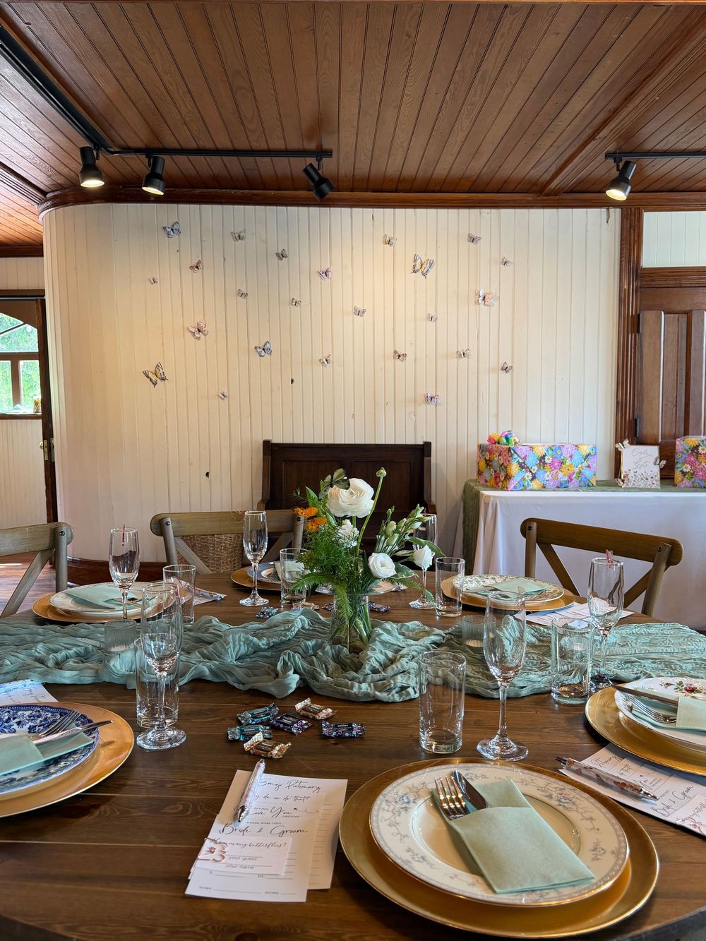 Elegant dining table set with gold chargers, green napkins, floral centerpiece, and butterfly wall decor in a wood-paneled room.