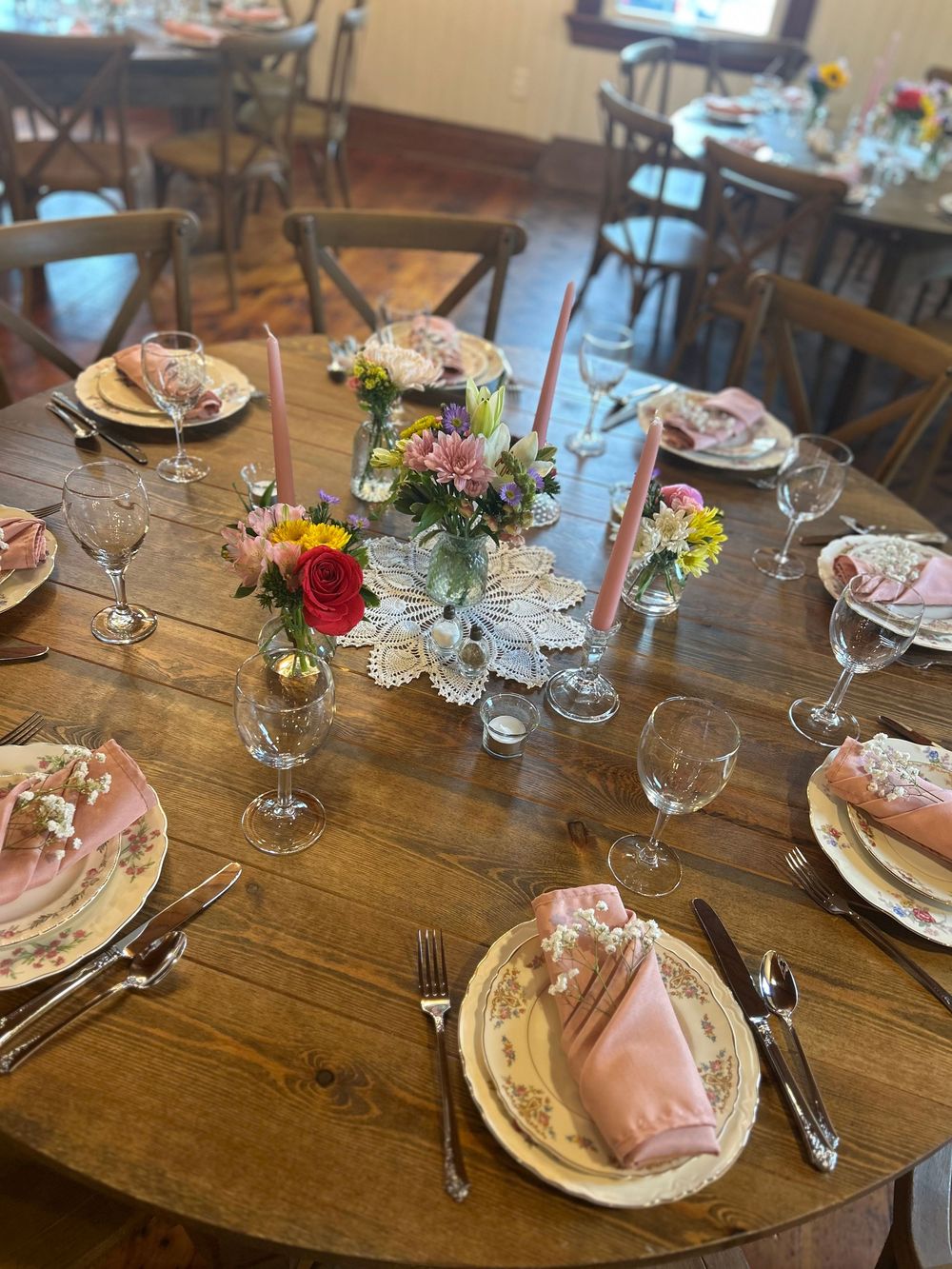 Elegant round wooden table set with vintage floral china, pink napkins, crystal glasses, and colorful flower centerpieces.