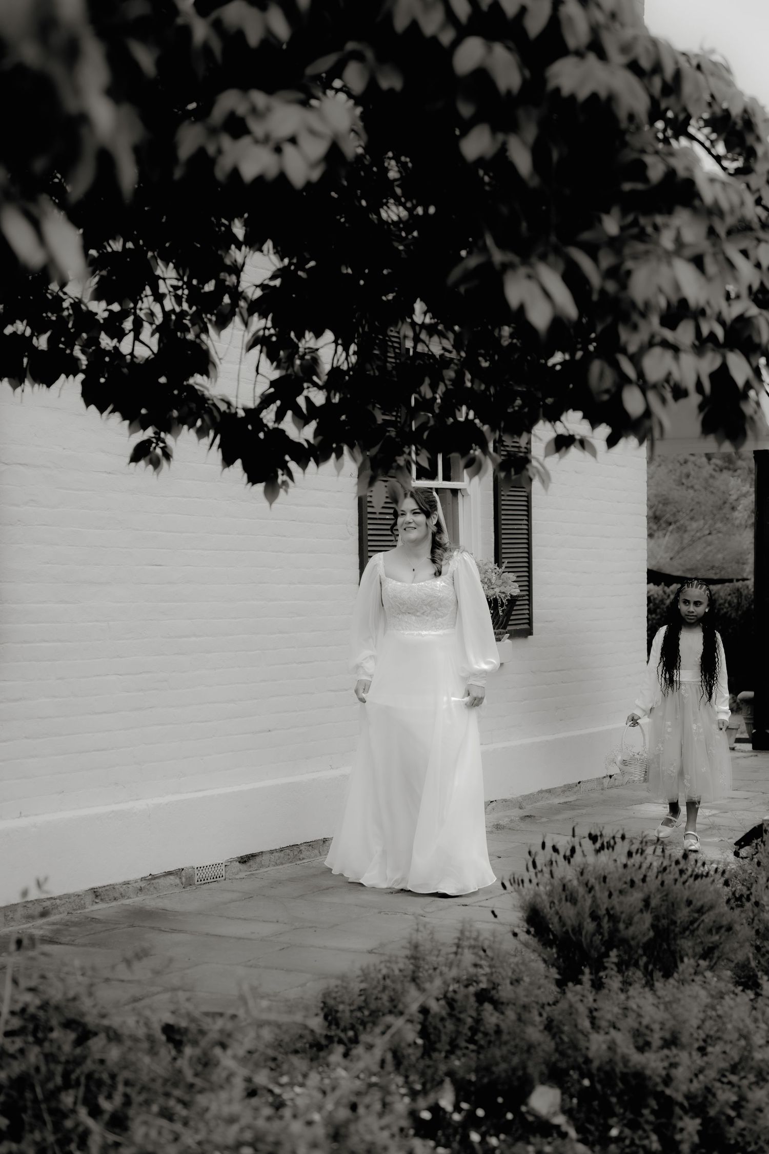 Black and white photo of a bride in a long white gown standing outside a white brick building with a child nearby.