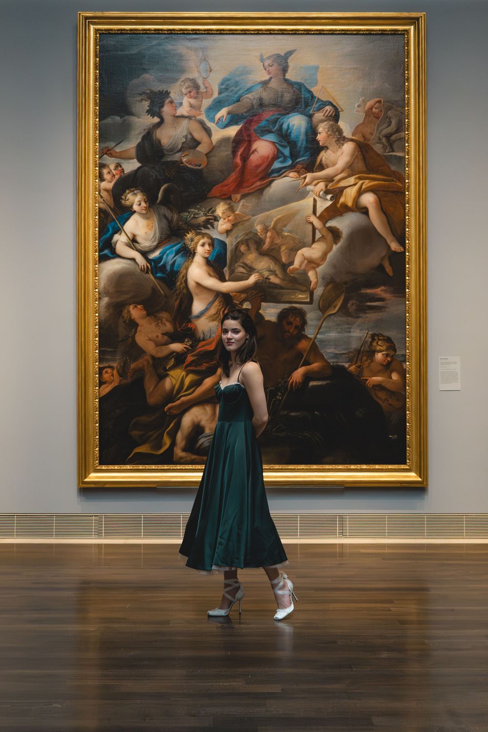 Young woman in green dress posing before large classical oil painting in art museum.