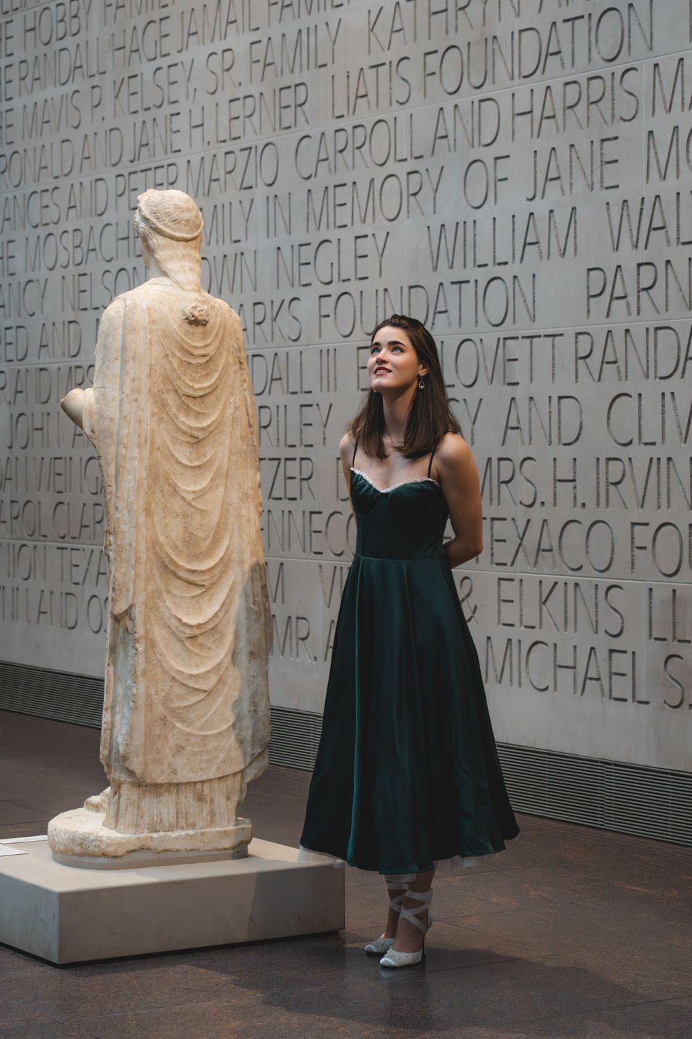 Elegant woman in green dress standing beside ancient marble statue in museum gallery.