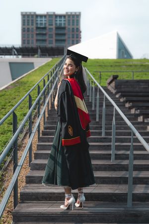 Graduate in cap and gown smiling on outdoor stairs at modern university campus.