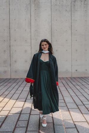Woman in green graduation gown and dress standing confidently against concrete wall.