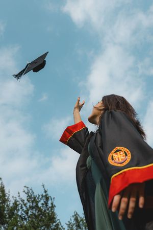 Graduate in black gown tosses mortarboard cap into blue sky celebrating academic achievement outdoors.