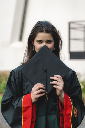 Female graduate in green gown holds black mortarboard cap covering her face outdoors.