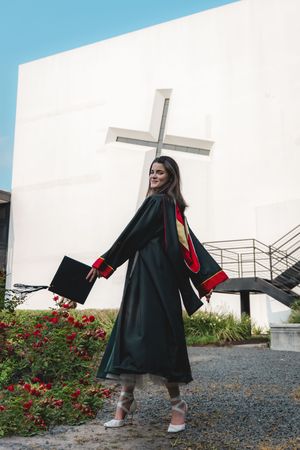 Smiling female graduate in green gown poses with arms spread near white cross monument.