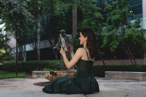 Young woman in green dress sits on ground photographing bronze sculpture in urban park.