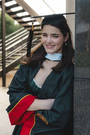 Smiling female graduate in green cap and gown leans against pillar with arms crossed.
