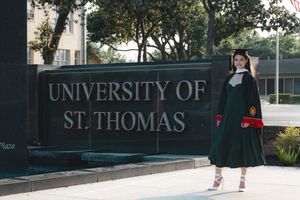 Female graduate in green cap and gown poses by University of St. Thomas entrance sign.