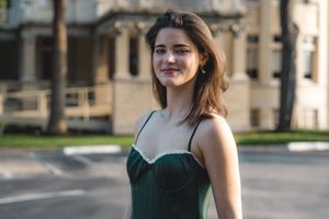 Young woman in green spaghetti strap dress smiles outdoors on sunny tree-lined street.