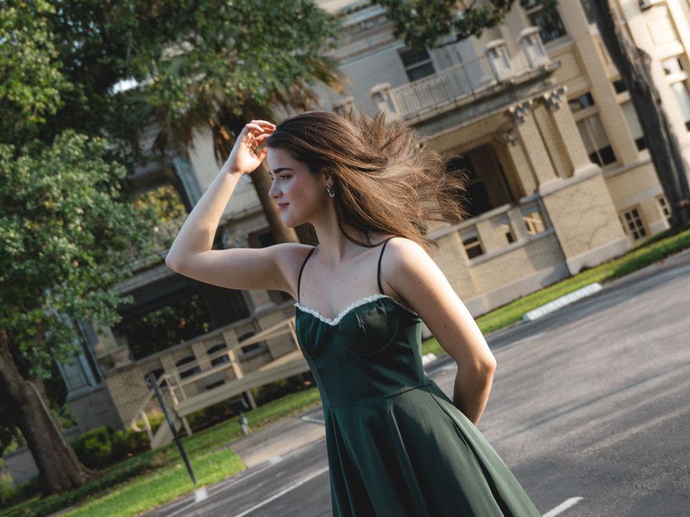 Young woman in flowing green dress with hair blowing in wind on sunny street.