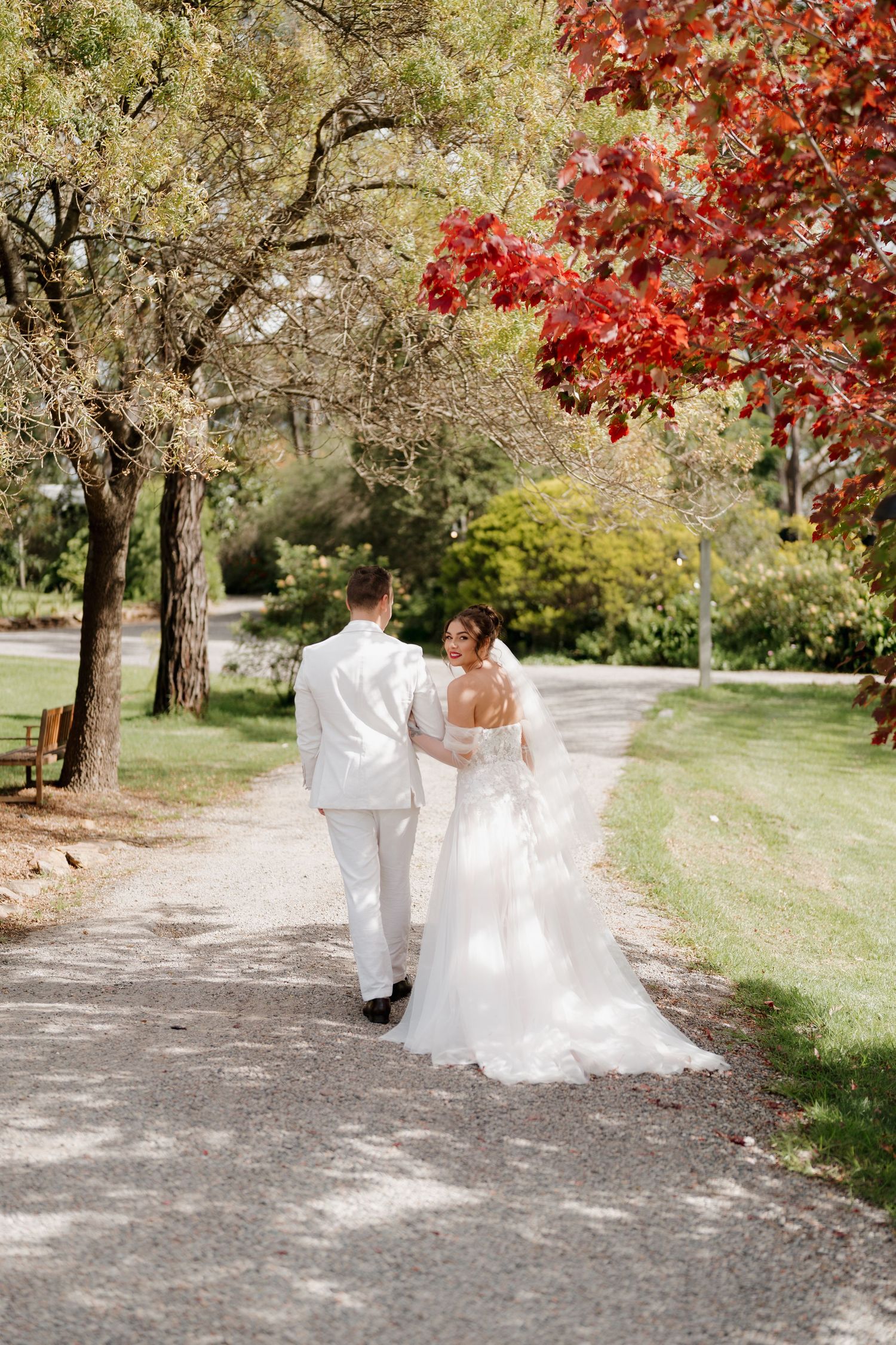Bride and groom walking hand in hand down a tree-lined path surrounded by lush green and red autumn foliage.