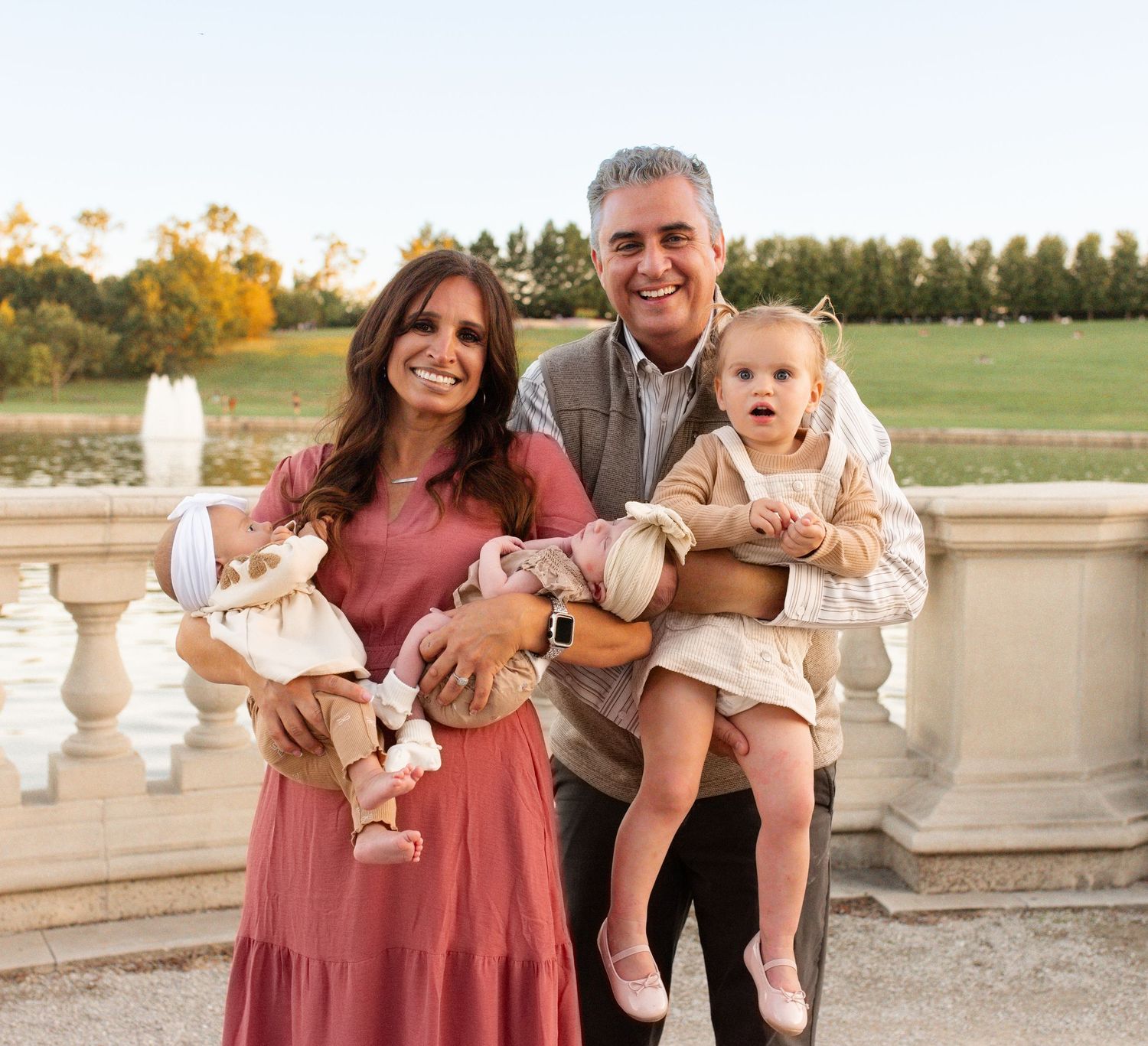 A smiling family poses at forest park, parents holding two young toddler girls in pink dresses near a scenic waterfront.