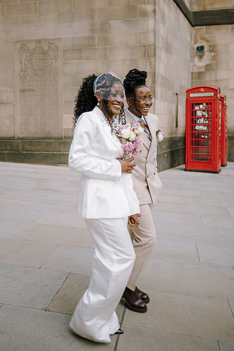 Two brides in white suits holding flowers outside a London building with a red telephone box.