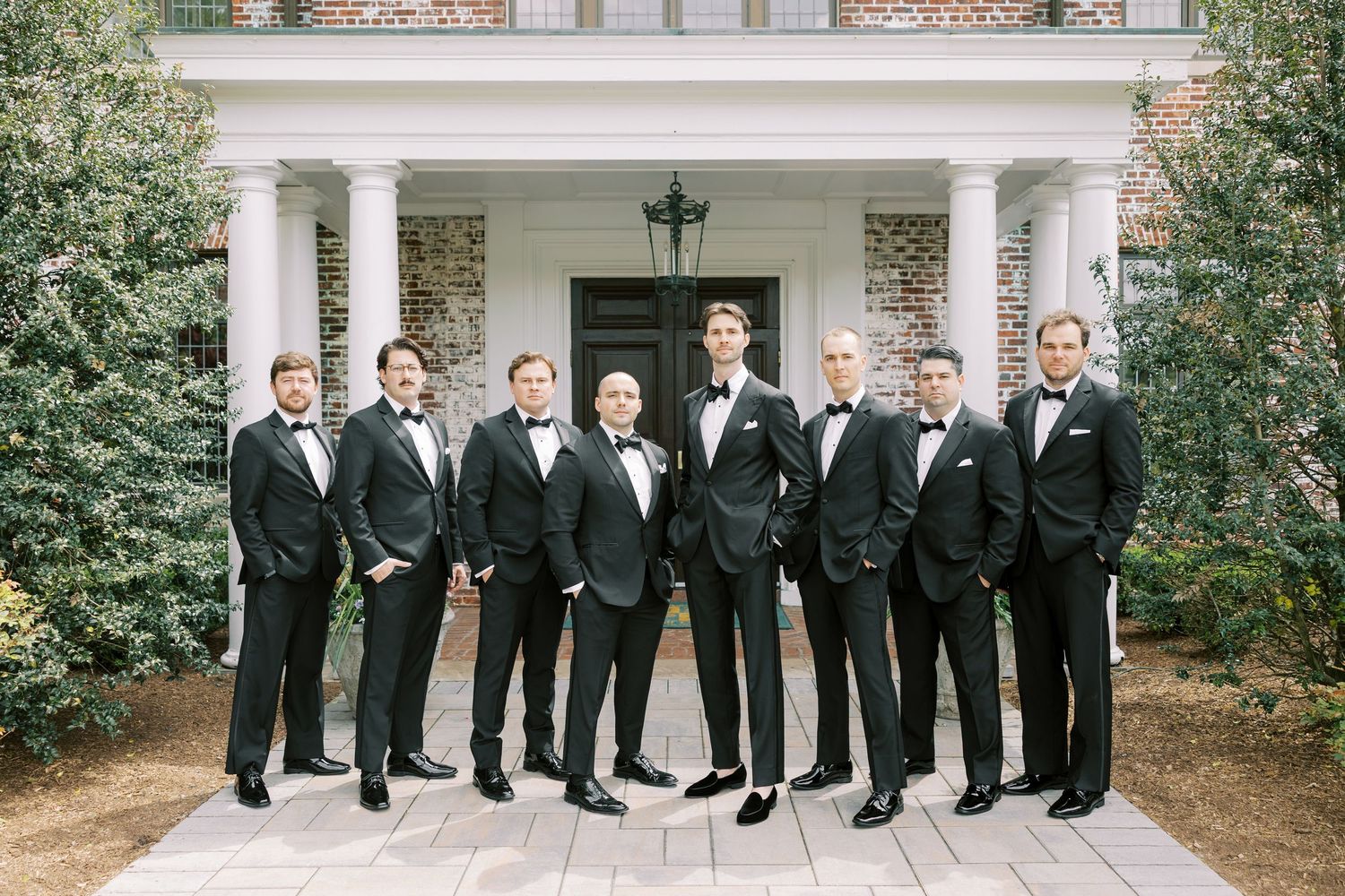Groom and groomsmen in black tuxedos with bow ties posing in front of elegant white columned mansion.