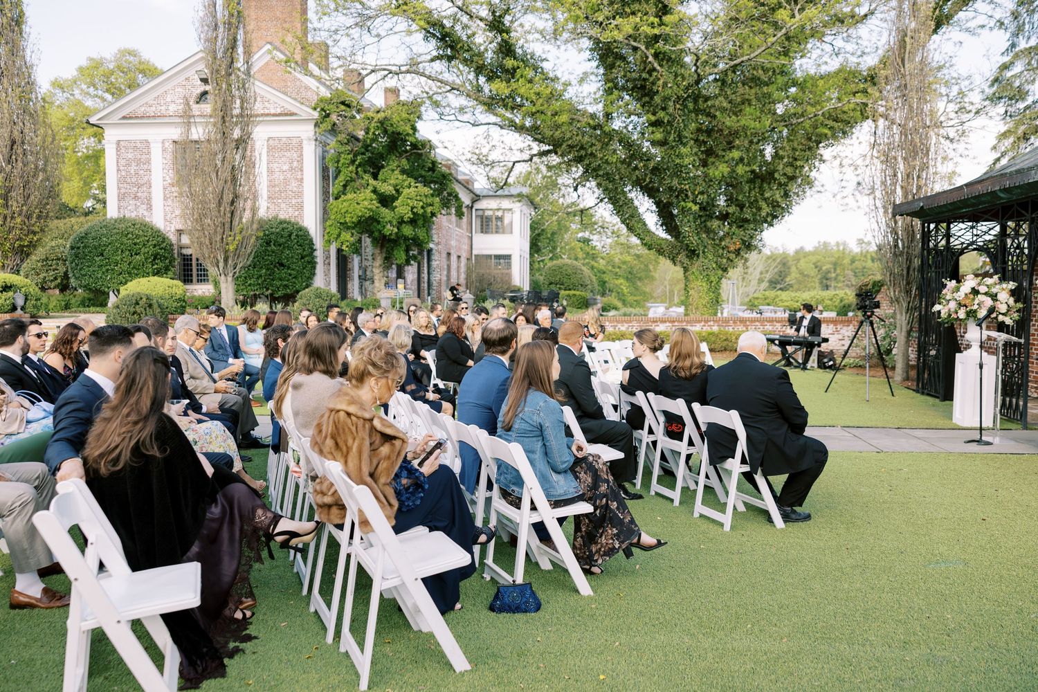 Guests seated in white folding chairs at an elegant outdoor wedding ceremony on a lush green lawn beside a historic estate.