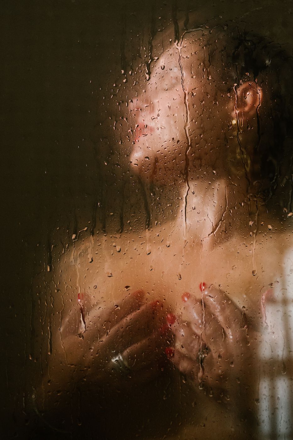 A person showering behind a wet glass door with water droplets, captured in warm, moody tones.