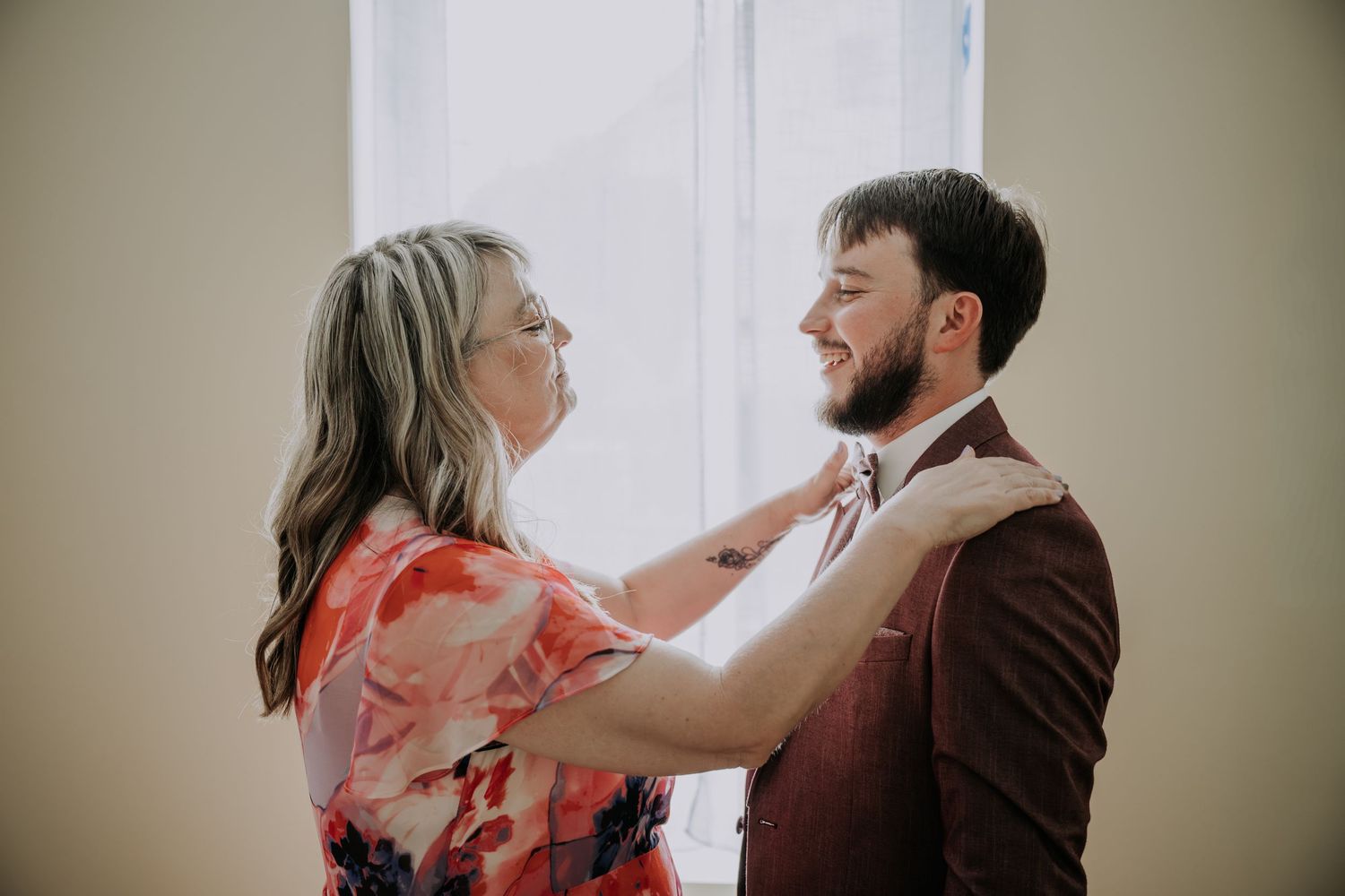 A woman in a floral dress adjusts a mans bow tie as he smiles, preparing for a wedding ceremony indoors.
