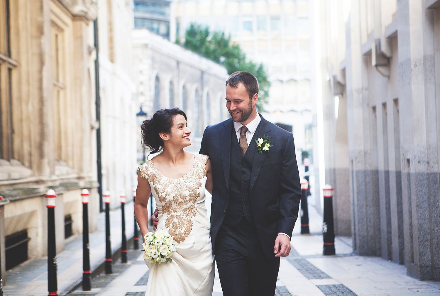 Smiling bride and groom walking together in an urban alley on their wedding day, bride holding a white bouquet.