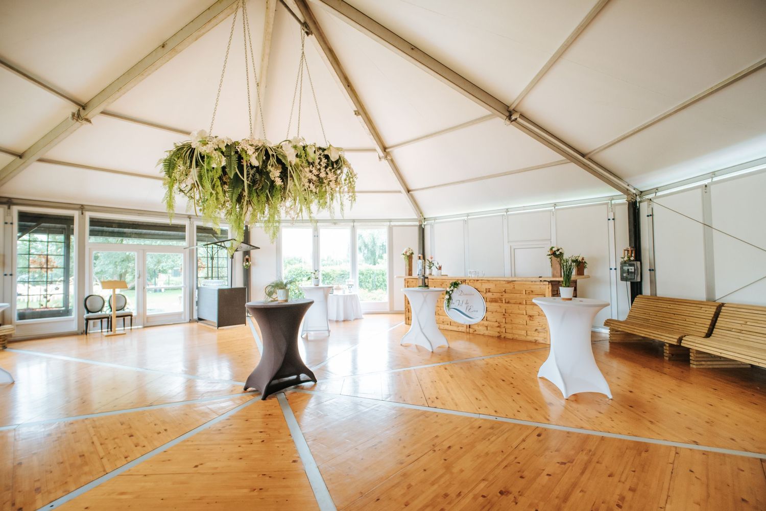Elegant tent venue with wood floors, high-top tables, hanging greenery chandelier, and natural light for events.