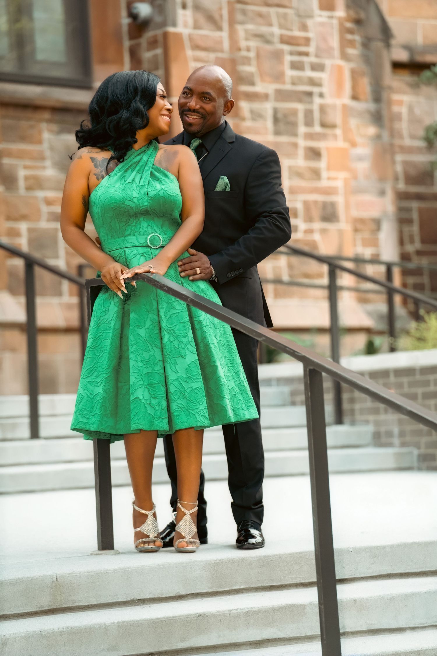 A couple poses on stone steps, woman in green halter dress, man in black suit with green pocket square.