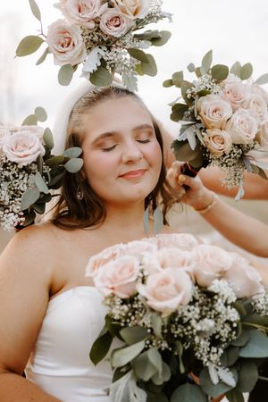 Bride in white dress with closed eyes surrounded by bouquets of blush pink roses and eucalyptus greenery.