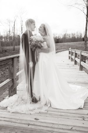 Black and white photo of bride and groom sharing a romantic moment under veil on rustic wooden bridge.