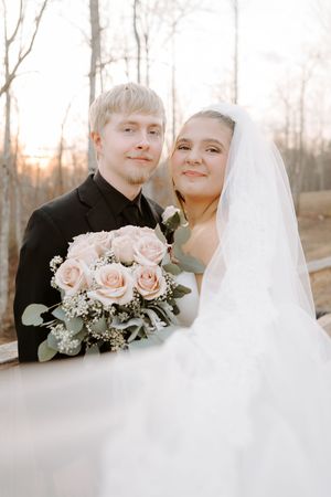 Bride and groom pose together on a wooden bridge holding a blush pink rose bouquet at golden hour.