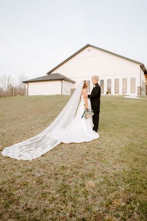 Bride and groom share romantic moment outdoors, bride's cathedral veil flowing beside white barn.