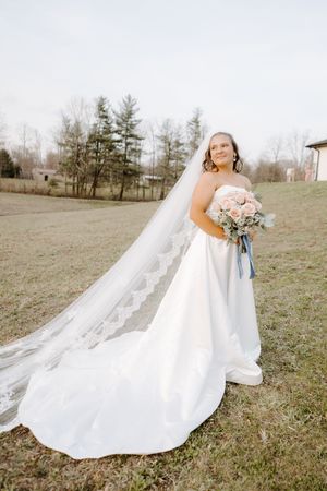 Bride in a strapless white gown with a long lace veil holds a pink bouquet in an outdoor field setting.