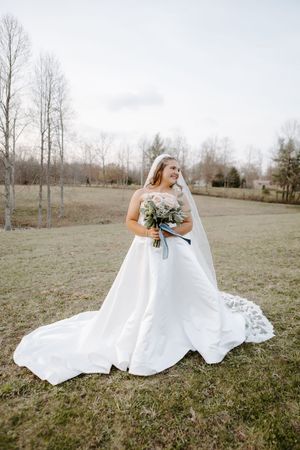 Bride in white ball gown with long train and veil holds bouquet outdoors on green grass with bare trees.