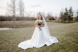 Bride in a classic white ball gown with long veil holding a bouquet on a green lawn.