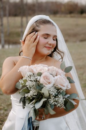 Bride in strapless gown holds blush rose bouquet with eyes closed, wearing veil outdoors at sunset.