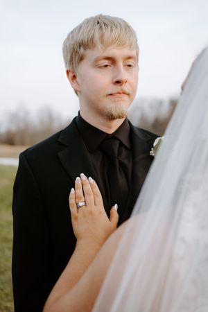 Groom in black suit with bride's hand showing diamond ring resting on his chest during outdoor wedding ceremony.